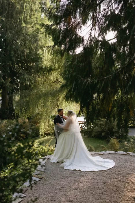Newlyweds embrace in the gardens at Chateau Lill, framed by towering trees and soft natural light for a romantic outdoor portrait.
