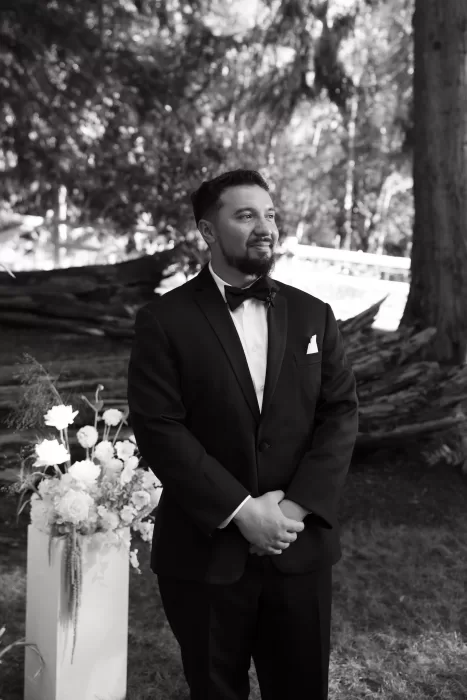 Black and white portrait of the groom waiting at the forest ceremony site at Chateau Lill, surrounded by floral arrangements.