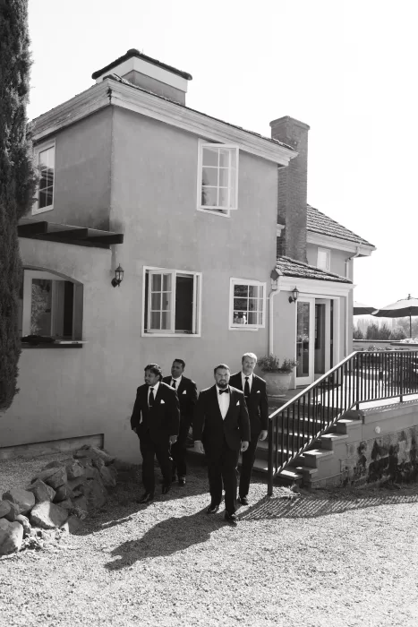 Groom and groomsmen walk together outside Chateau Lill’s villa buildings, dressed in classic black tuxedos before the ceremony.
