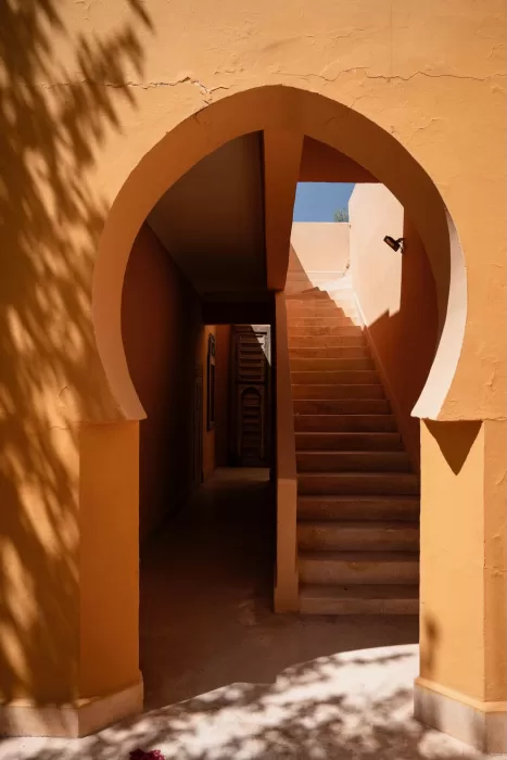 Warm terracotta arch framing a sunlit stairway at Jnane Tamsna, with soft shadow patterns on the walls.