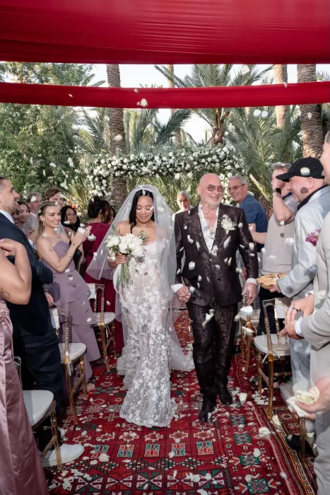 Newly married couple walking the aisle under red canopy fabric at Jnane Tamsna as guests toss white rose petals.