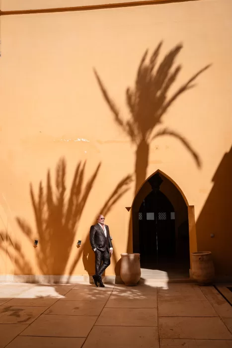 Groom standing beneath dramatic palm shadows cast across an ochre courtyard wall at Jnane Tamsna.