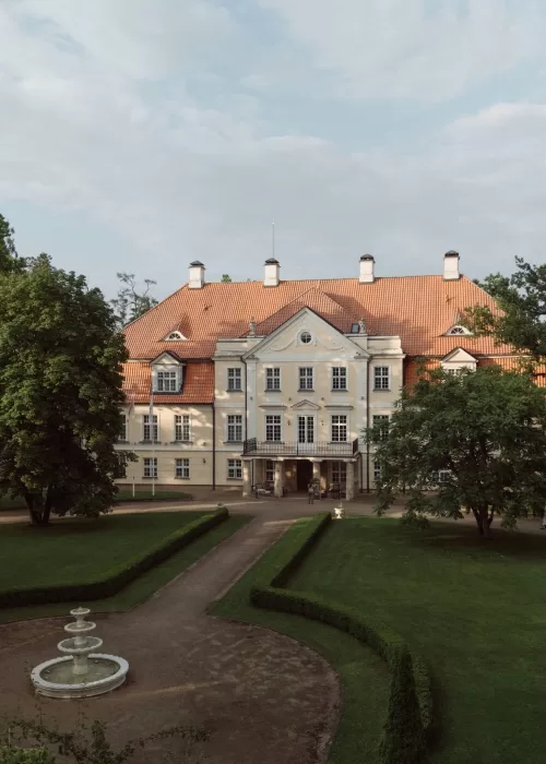 Elevated view of Malpils Manor revealing its grand symmetrical facade, formal gardens and central fountain in soft daylight.