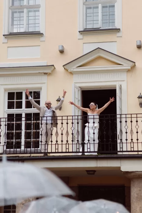 Bride and groom celebrating on a balcony at Malpils Manor, arms raised as guests gather below.