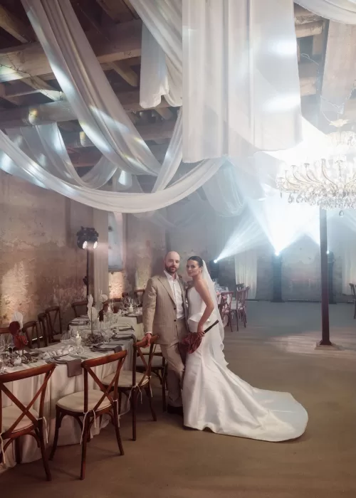 Bride and groom standing together inside the Malpils Manor reception space beneath soft draped fabric and warm lighting.