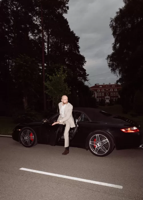 Groom stepping out of a black sports car near Malpils Manor at dusk, creating a cinematic wedding arrival moment.
