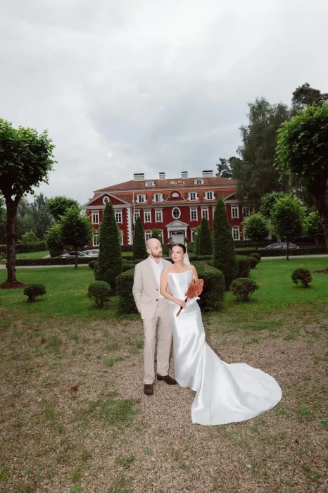 Bride and groom standing together in the formal gardens of Malpils Manor, with the historic red-brick facade behind them.
