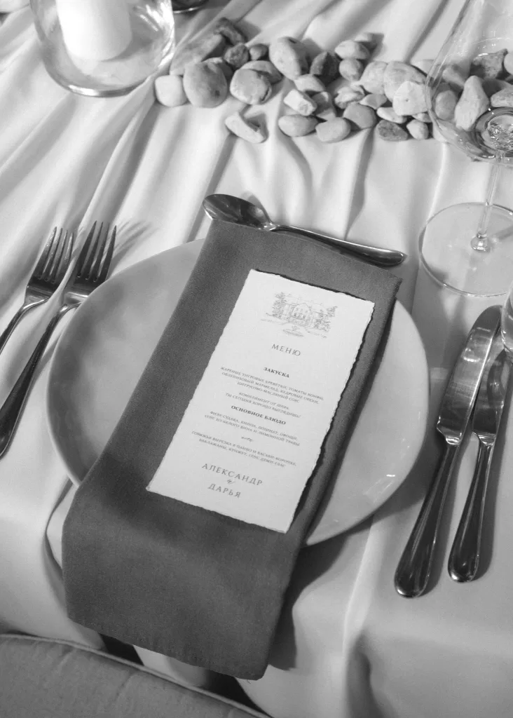 Close-up of a wedding place setting at Malpils Manor, featuring printed menu cards, stone accents and refined tableware.