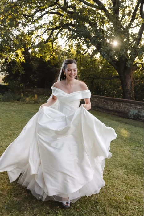 The bride lifts her gown playfully while walking across the sunlit lawn at Villa Lena.