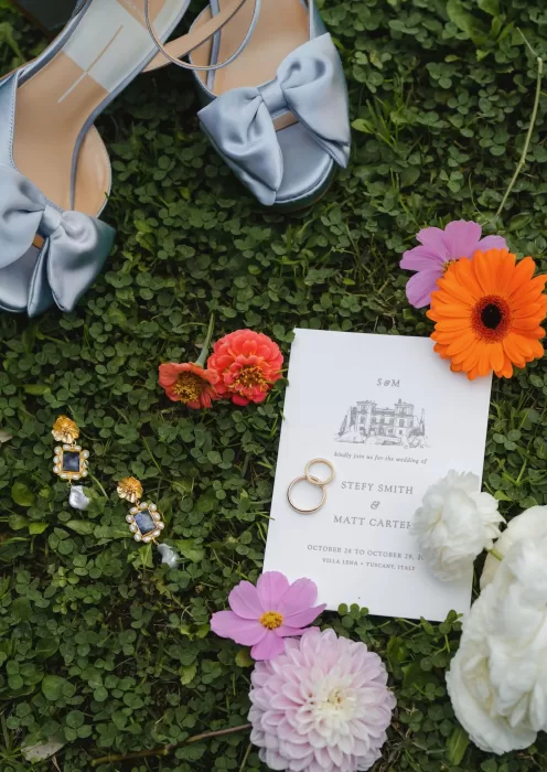 Wedding details arranged on grass at Villa Lena, including blue satin heels, jewellery, flowers, and the invitation suite.