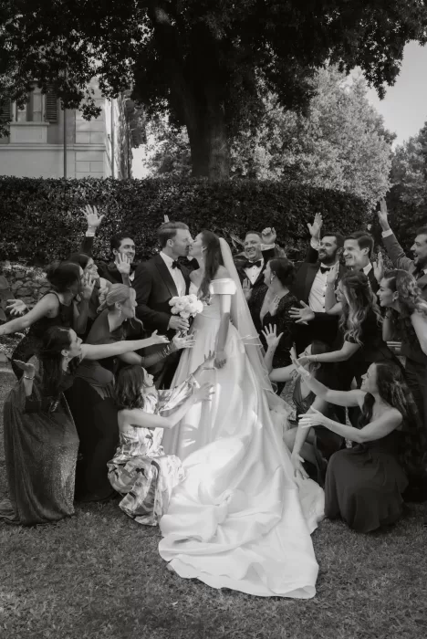 The couple kisses surrounded by cheering friends in an energetic group portrait on the Villa Lena lawn.