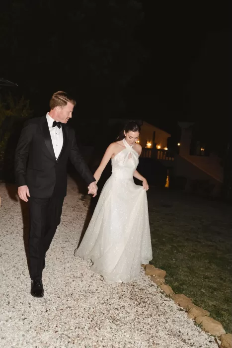 The couple walks hand in hand through the Villa Lena grounds at night, her glittering gown catching the warm lights.