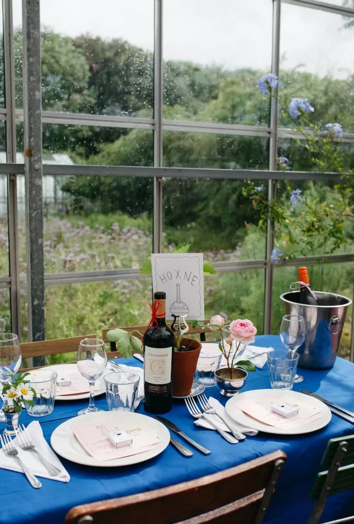 Blue-linen wedding table inside the greenhouse at Worton Kitchen Garden, set with flowers, wine, and illustrated table signage.