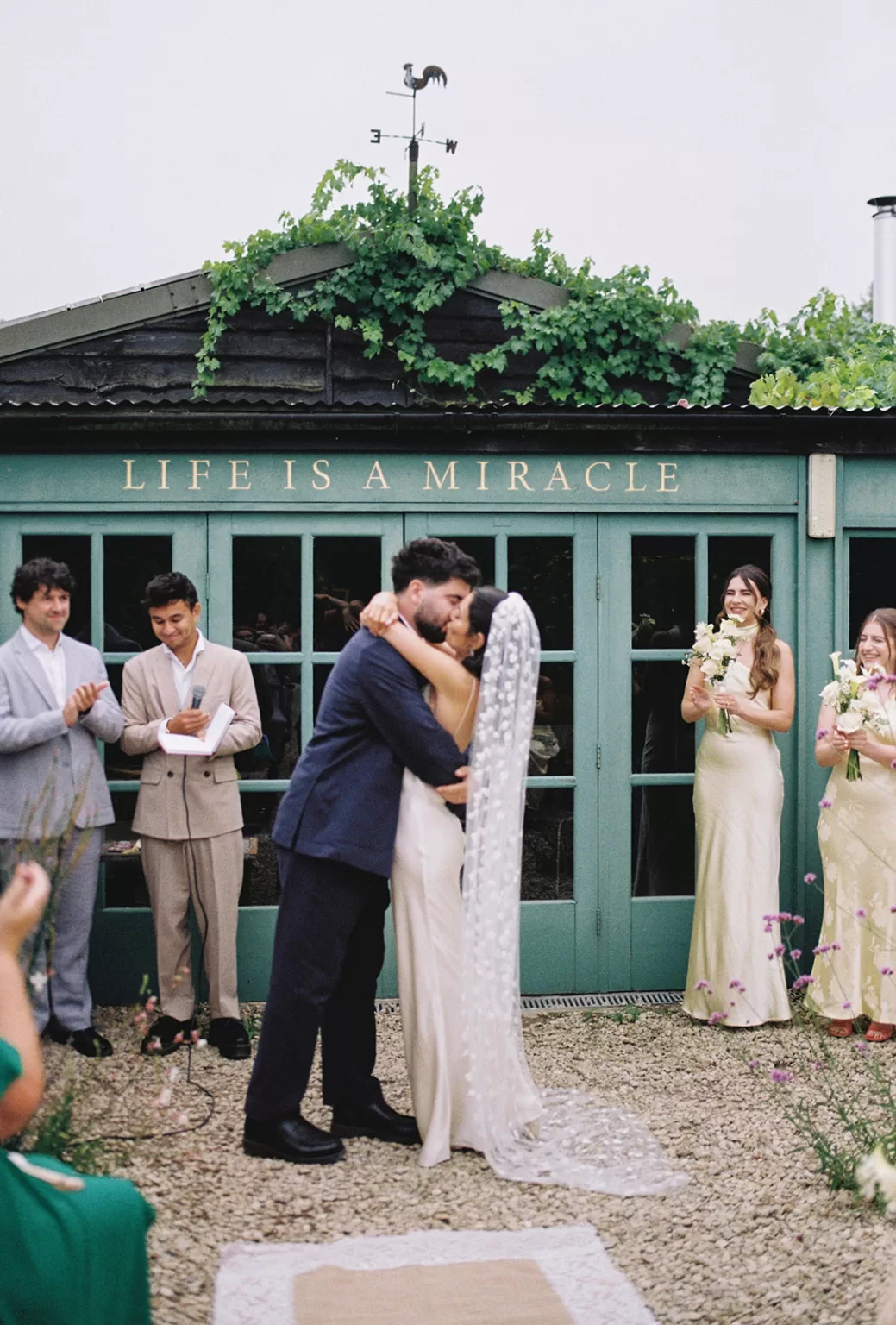 Couple sharing a kiss during their outdoor ceremony at Worton Kitchen Garden, surrounded by greenery and smiling guests.