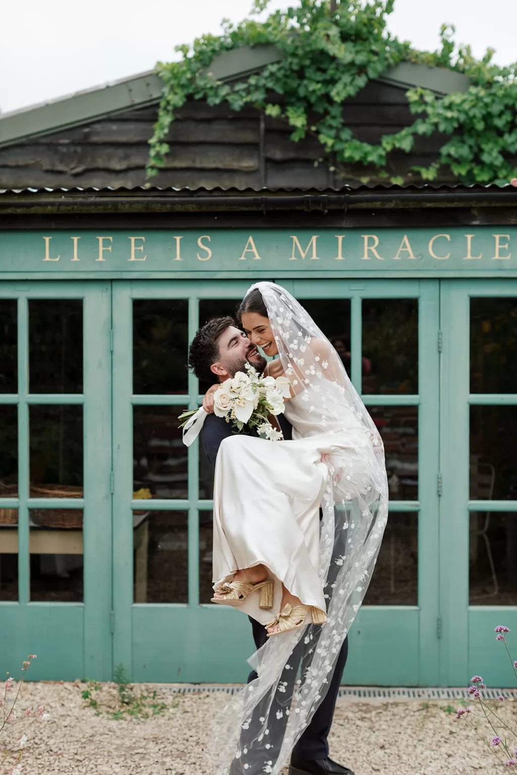 Groom lifting his bride playfully in front of the “Life Is a Miracle” building at Worton Kitchen Garden, her veil flowing behind them.