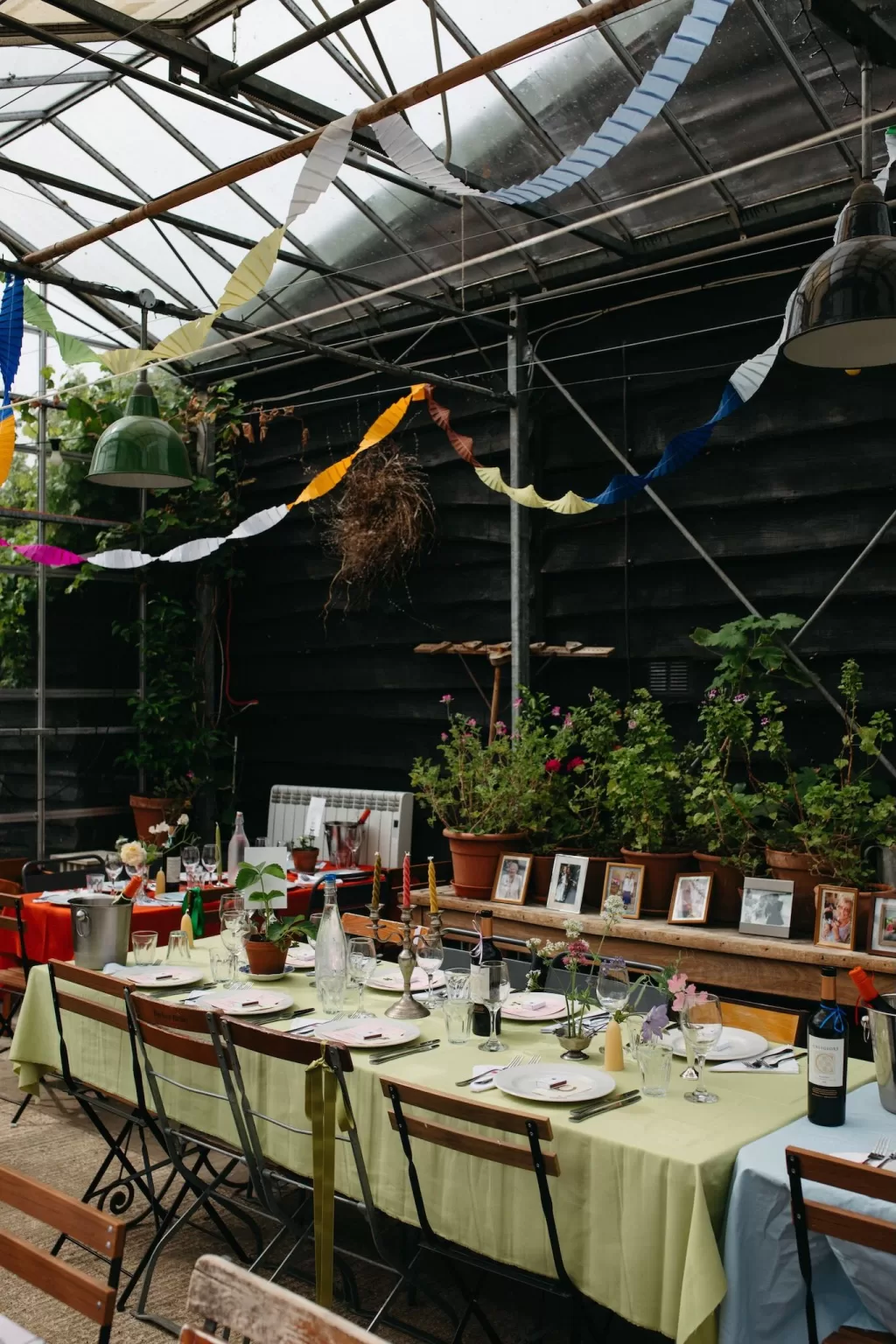 Colourful streamers and long dinner tables inside the Worton Kitchen Garden glasshouse, styled with potted plants and framed photos.