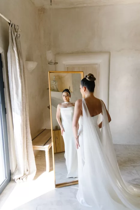 Bride standing in a softly lit suite at Cavo Ventus, wearing a minimalist wedding dress and veil while admiring her reflection.