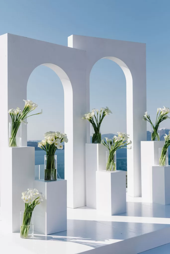 White ceremony arches and calla lily arrangements at Cavo Ventus, framed against clear blue skies and sea views.