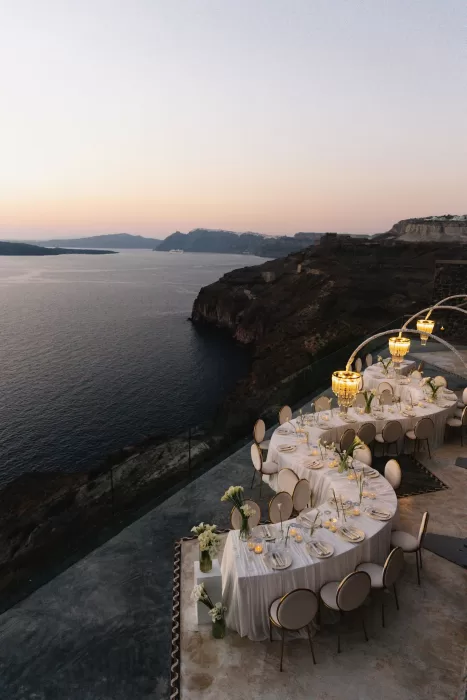 Curved wedding reception tables at Cavo Ventus set along the cliffs at dusk, softly lit with candles and chandeliers overlooking the Santorini caldera.