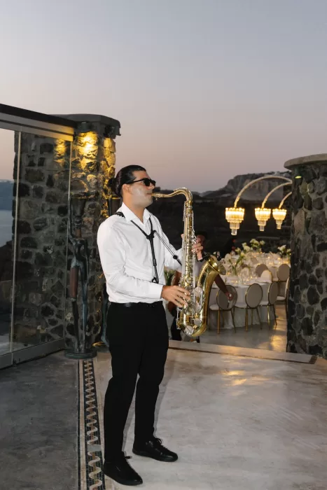 Saxophonist performing during an outdoor wedding reception at Cavo Ventus, with candlelit tables and stone architecture in the background.