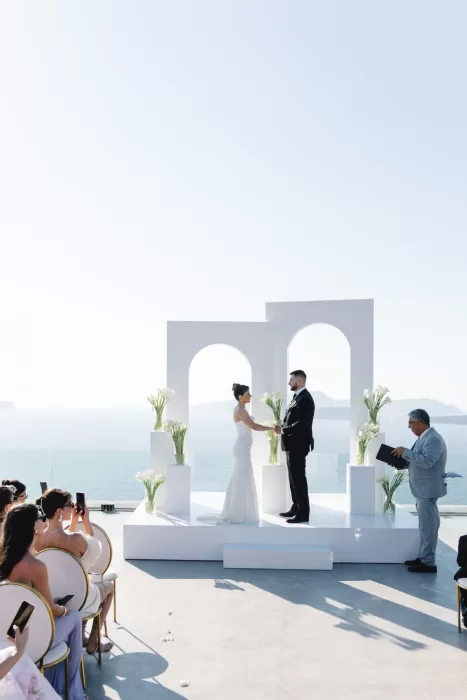 Modern wedding ceremony at Cavo Ventus with white architectural arches and floral pedestals set against panoramic Santorini sea views.