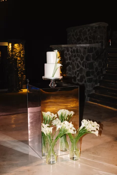 Minimalist white wedding cake displayed on a mirrored plinth at Cavo Ventus, styled with calla lilies and dramatic evening lighting.