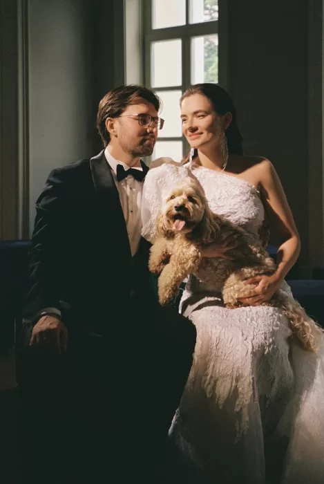 Bride and groom seated together holding their dog, photographed in soft natural light with a timeless editorial mood.