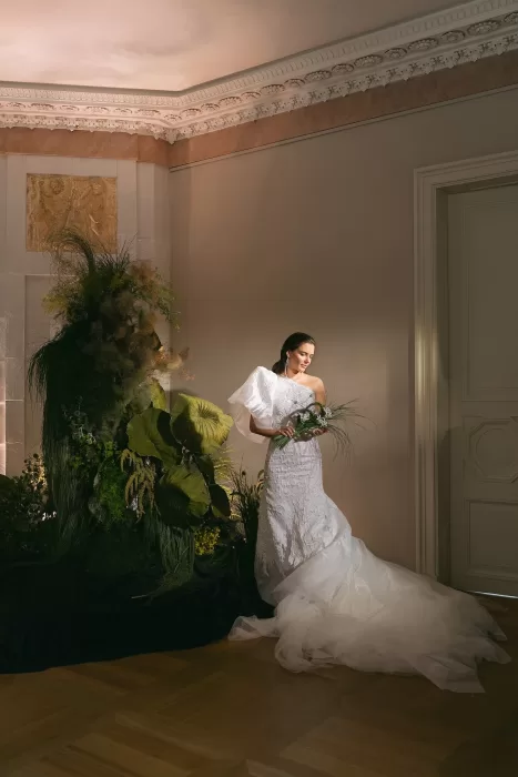 Bride standing beside a sculptural floral installation, wearing a textured one-shoulder gown in a softly lit editorial setting.
