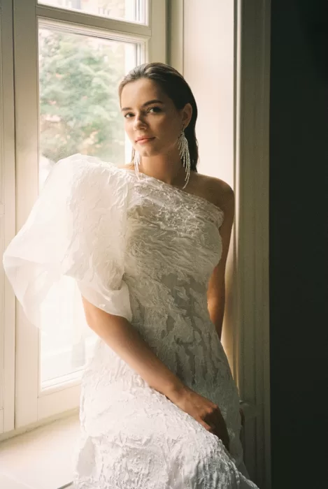 Editorial bridal portrait by a window, highlighting textured fabric, natural light, and a calm, refined mood.