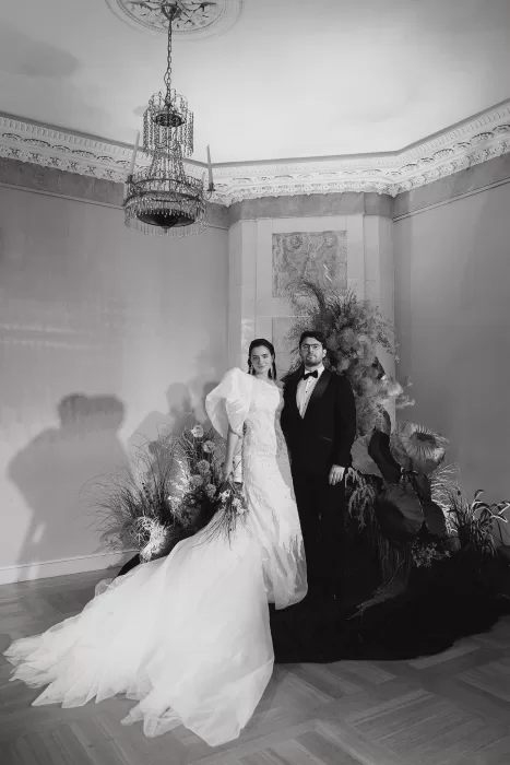 Black and white editorial portrait of a bride and groom standing beside a sculptural floral installation in a refined interior.