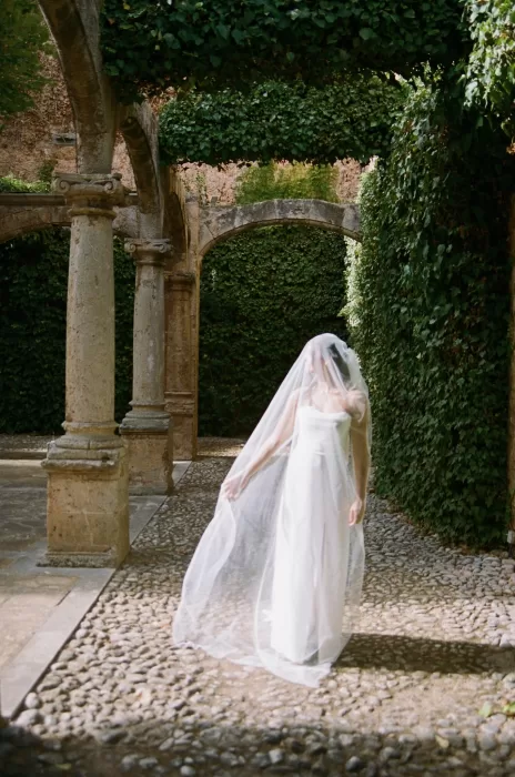 Bride walking beneath stone arches with her veil flowing through the cloister at Finca Es Convent.
