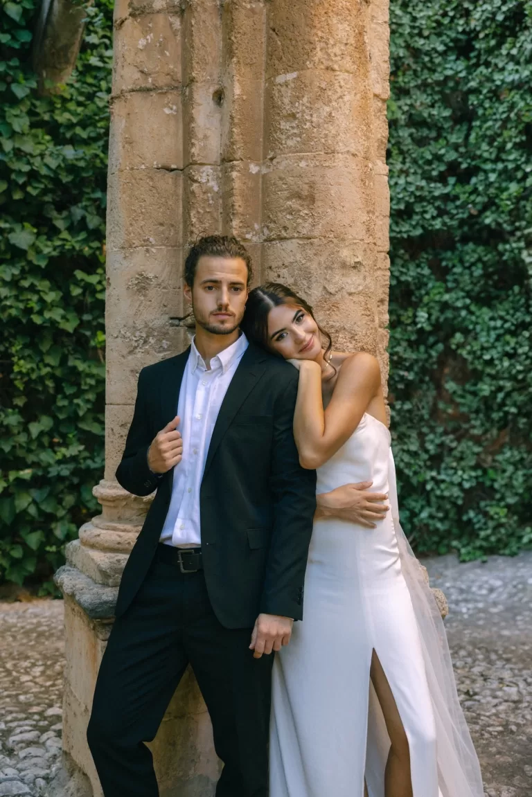 Bride leaning into her groom beside a historic stone column at Finca Es Convent, framed by ivy-covered walls.