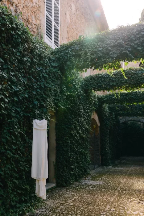 Minimal wedding dress hanging in an ivy-lined stone walkway at Finca Es Convent, bathed in natural light.