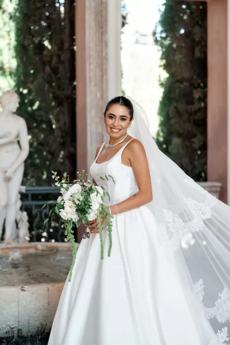Bride smiling beside a stone fountain at Finca La Concepción, wearing a flowing veil and holding a soft white bouquet.