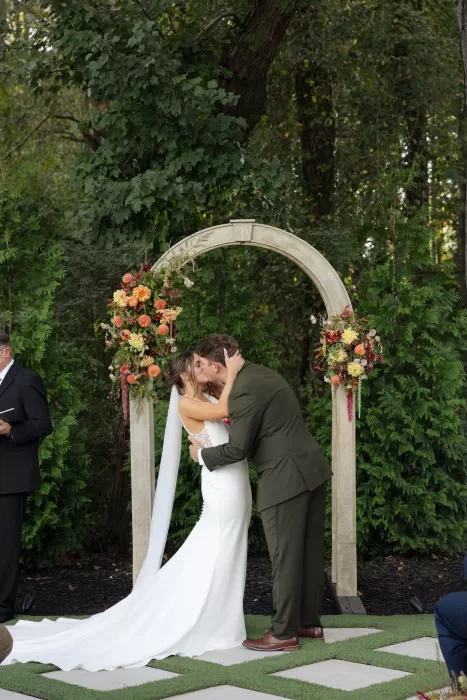 The couple share their first kiss beneath the ceremony arch at The Greenhouse at Arrows Farm, surrounded by autumn florals.