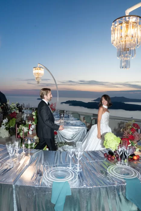 Bride and groom walking between reflective reception tables at sunset at Islet Santorini, overlooking the caldera.