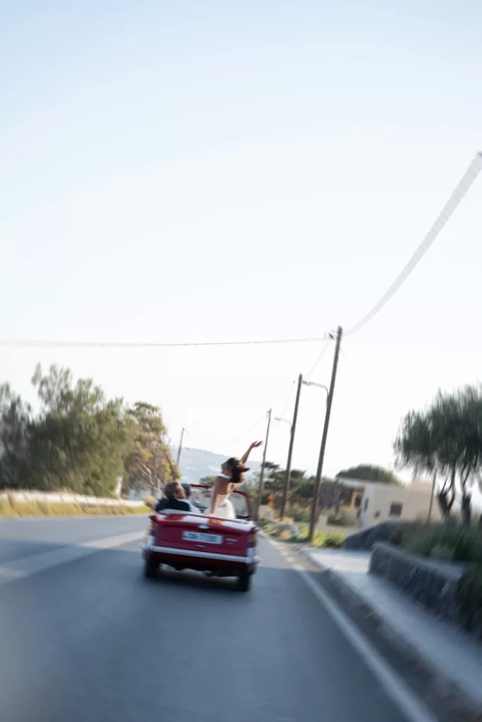 Bride and groom driving away in a red vintage convertible after their wedding at Islet Santorini.
