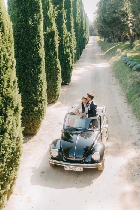 Bride and groom riding in a vintage convertible along the cypress-lined driveway at Mas de Les Voltes.