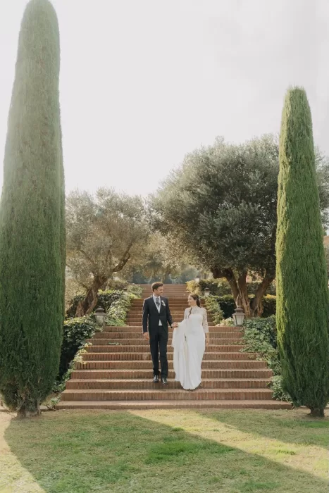 Bride and groom descending the garden steps at Mas de Les Voltes, framed by olive trees and cypress-lined paths.