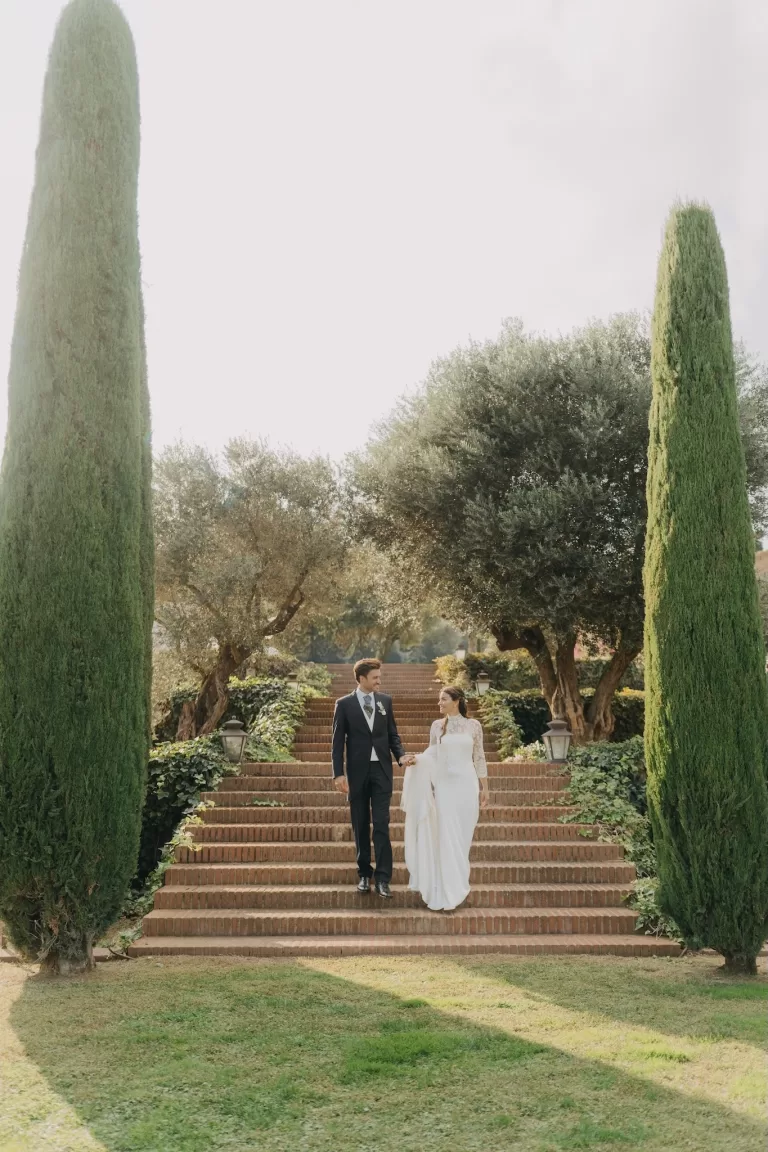 Bride and groom descending the garden steps at Mas de Les Voltes, framed by olive trees and cypress-lined paths.