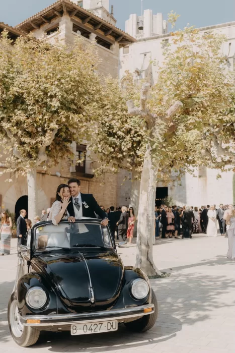Bride and groom waving from a vintage car on arrival at Mas de Les Voltes, with guests gathered outside the stone building.