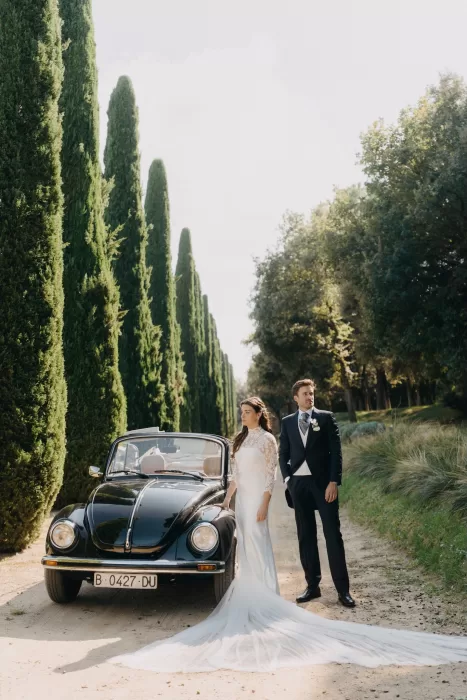 Bride and groom posing with a classic black car along the cypress-lined driveway at Mas de Les Voltes, surrounded by soft afternoon light.