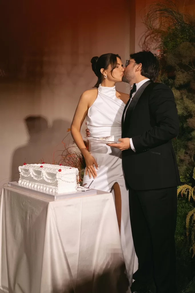 Bride and groom sharing a kiss beside a minimalist wedding cake, captured in warm evening light with an editorial feel.