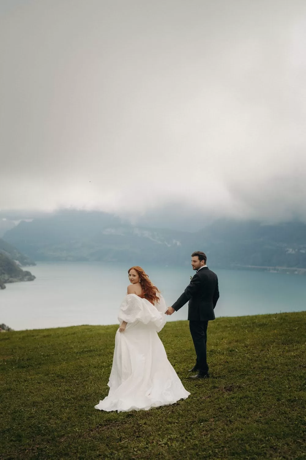 Bride turns to the camera as she and the groom walk hand in hand overlooking the lake and misty mountains at Hotel Villa Honegg.