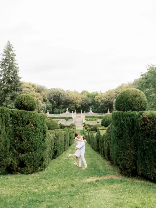 Couple embracing in the formal hedge gardens at Castello di Celsa with symmetrical pathways and stone staircase beyond.