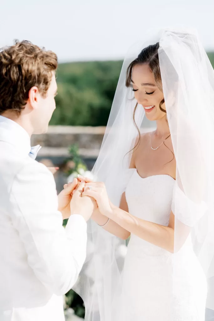 Bride smiling beneath her veil as groom places wedding ring during outdoor ceremony at Castello di Celsa.