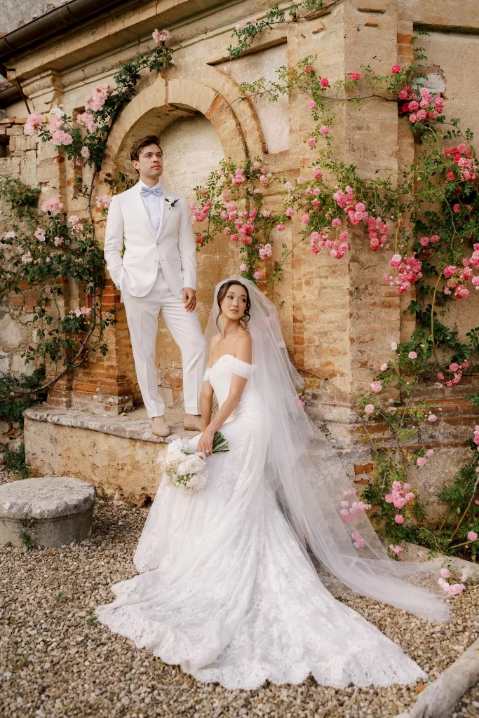 Bride and groom posing against climbing pink roses on a historic stone wall at Castello di Celsa in Tuscany.