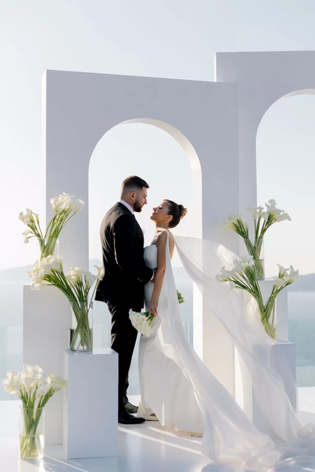 Bride and groom standing beneath white ceremony arches at Cavo Ventus, with flowing veil and calla lilies against the Aegean Sea.
