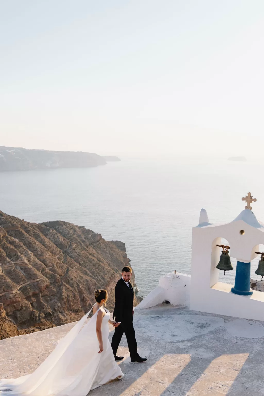 Bride and groom walking along the cliffs near Cavo Ventus with sweeping views of the Santorini caldera and Aegean Sea.