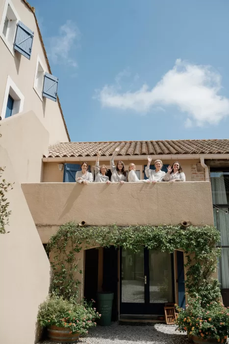 Bridesmaids waving from a sunlit balcony at Château Canet, framed by blue shutters and climbing greenery against warm stone walls.
